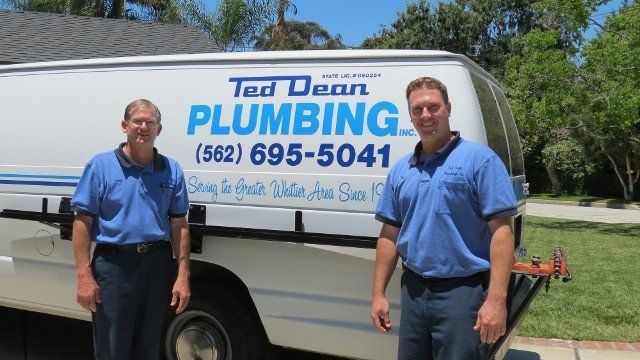 Two Ted Dean Plumbing techs in blue work shirts standing in front of the white company van, lettered with the phone number (562) 695-5041